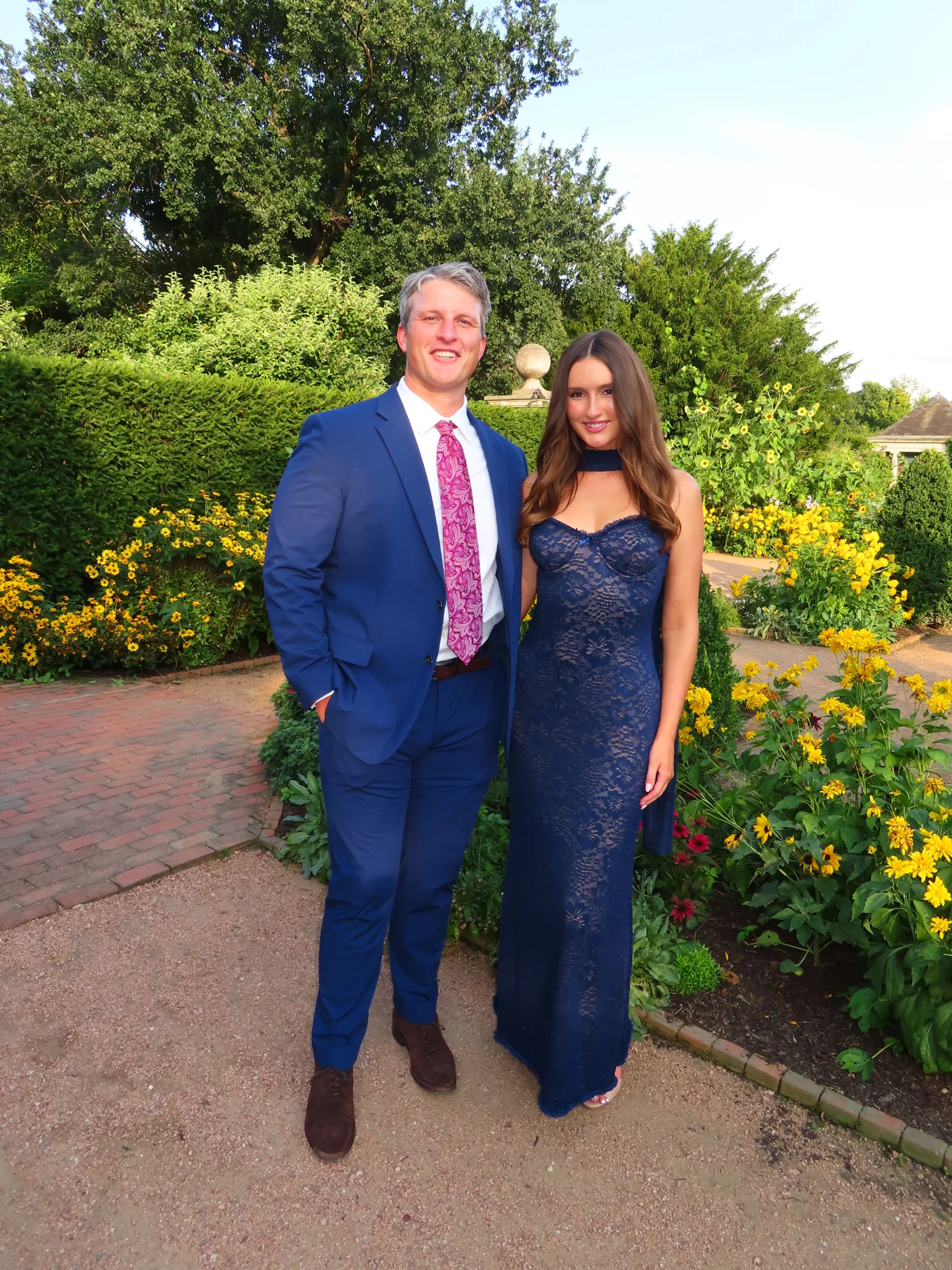 Noah in a blue suit with his girlfriend in a blue dress, standing together in a garden.